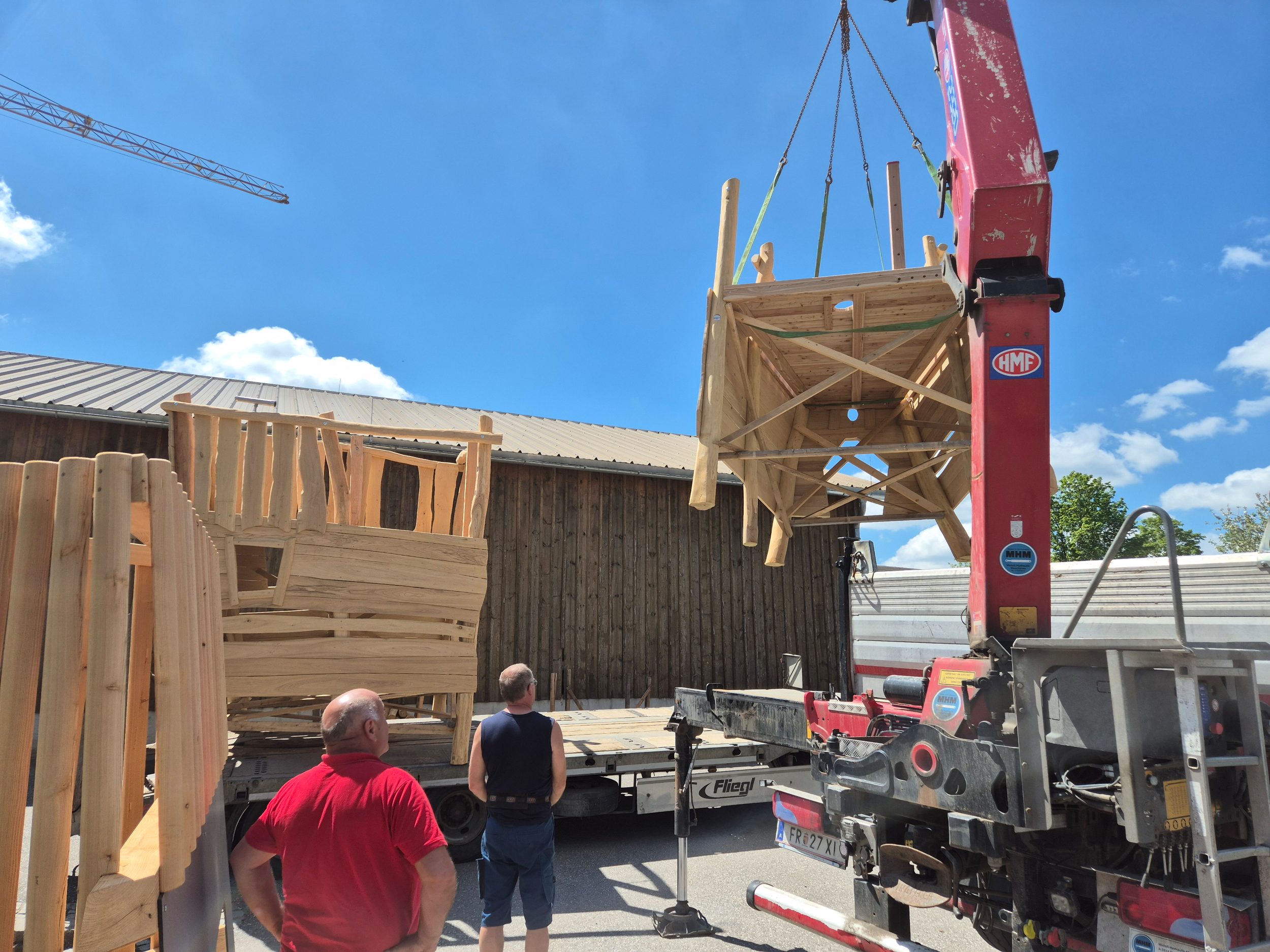 Kran hebt ein vorgefertigtes Holzelement vom Lkw, zwei Mitarbeiter begleiten die Verladung